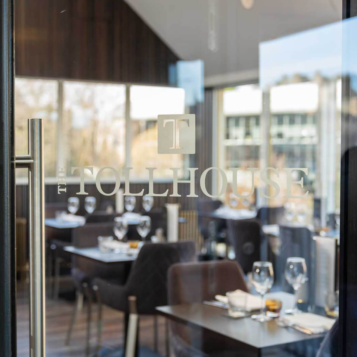 The Tollhouse etched on the glass, the bright dining room and trees along the riverbank beyond